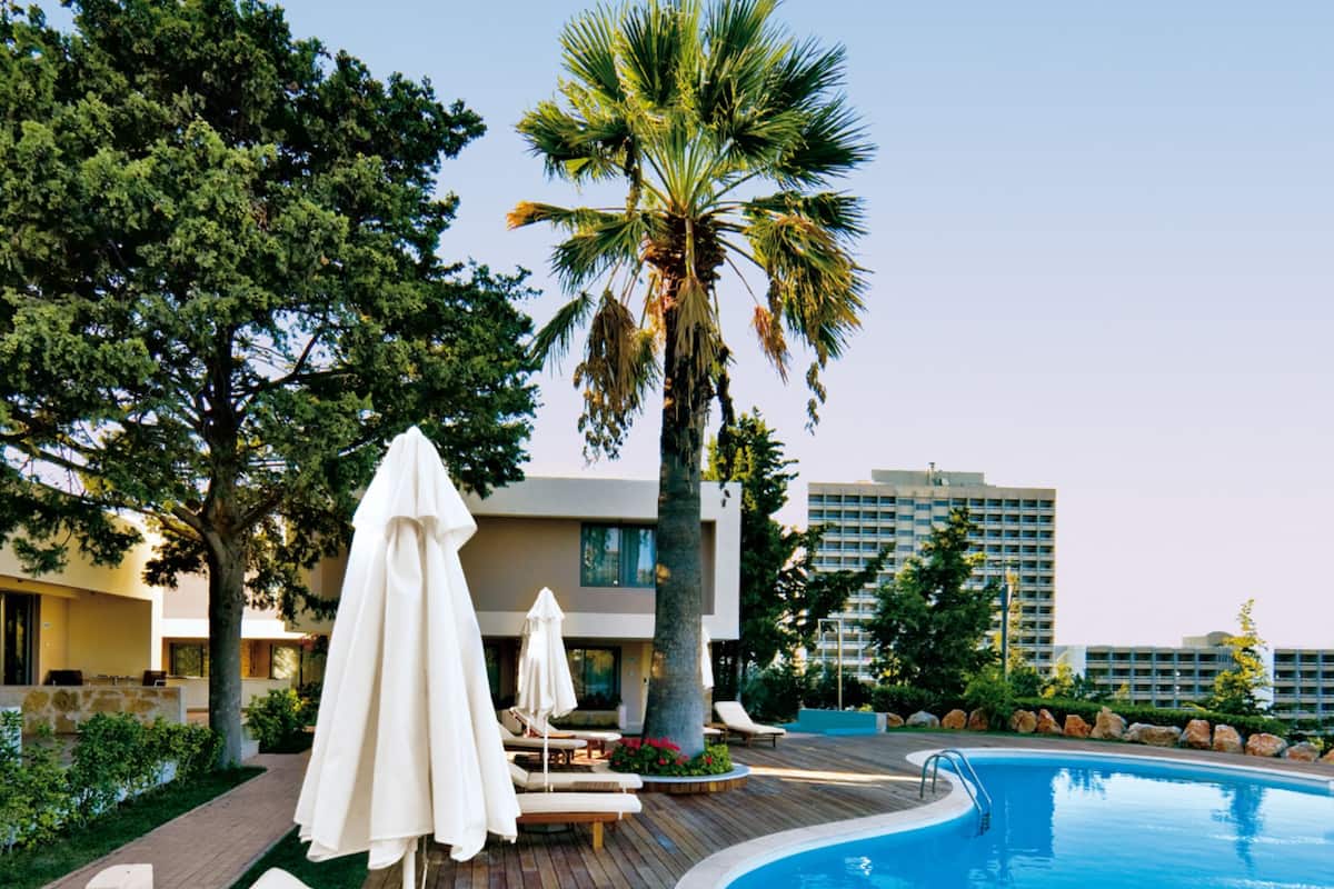 A poolside area with lounge chairs and umbrellas, surrounded by greenery and a modern building in the background.