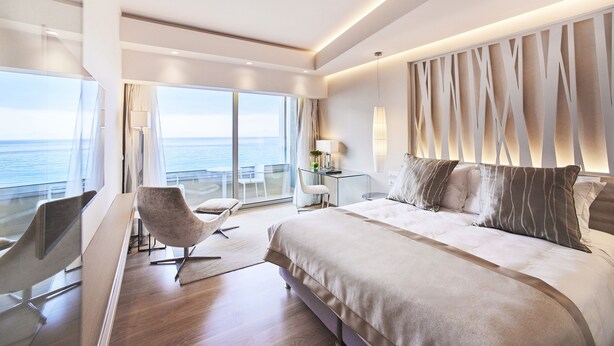 A modern hotel room with a large bed, a desk, and a chair. There is a view of the ocean through large windows. - Rodos Palace Hotel (Rhodes)