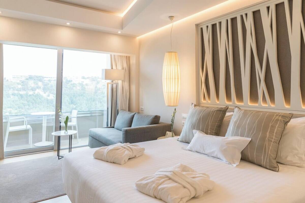 A modern hotel room with a large bed, a seating area with a sofa and armchairs, a balcony with a table and chairs, and a decorative wall panel.