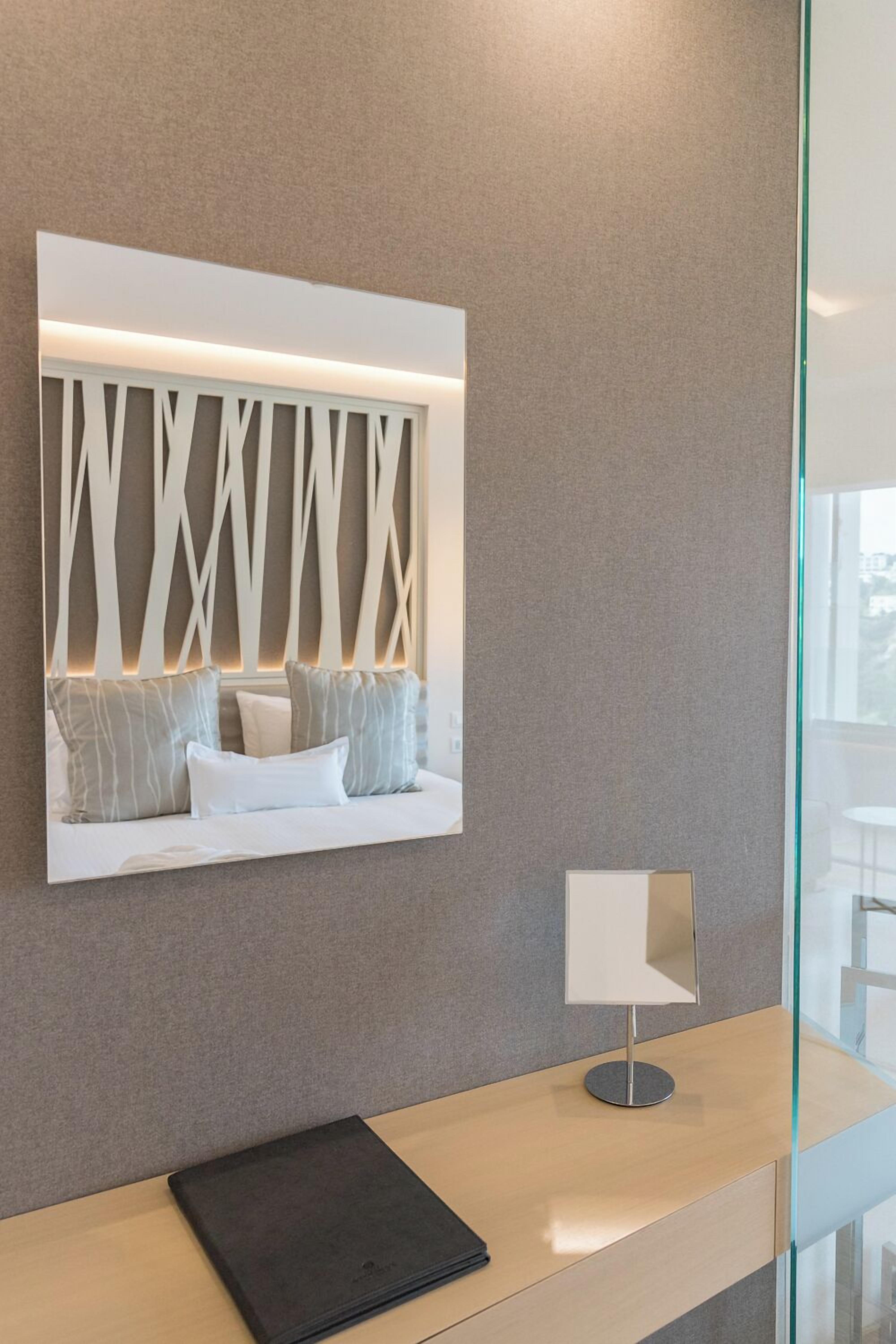 A modern hotel room with a grey textured wall, a white decorative panel with geometric patterns, a sofa with grey and white cushions, a small table with a lamp, and a black folder on the table.