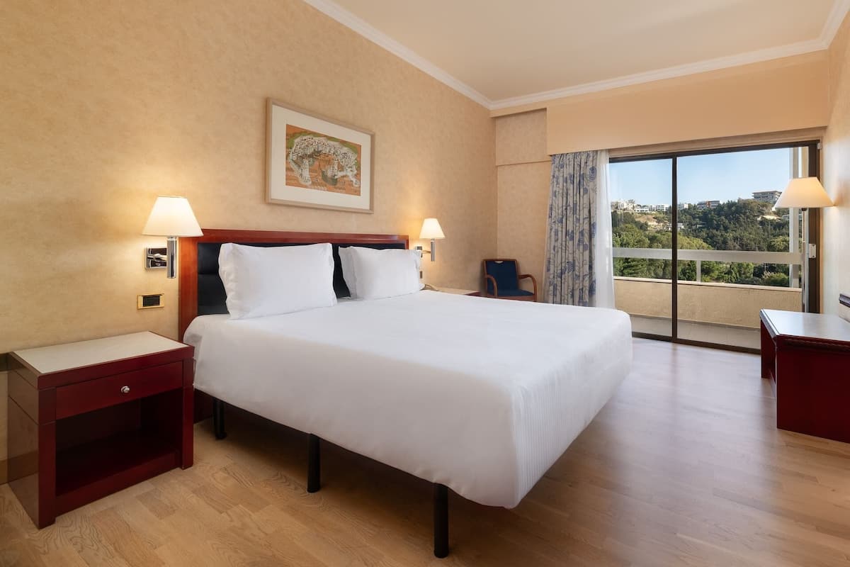 A hotel room with a large bed, two bedside tables, a chair, a window with a view, and a painting on the wall.