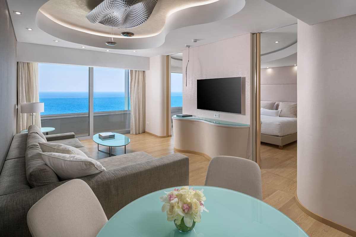 A modern living room with a sofa, a glass table with a vase, a flat-screen TV, and a balcony with a view of the sea.