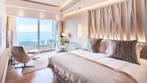 A modern hotel room with a large bed, a desk, a chair, and a view of the ocean. - Rodos Palace Hotel (Rhodes)