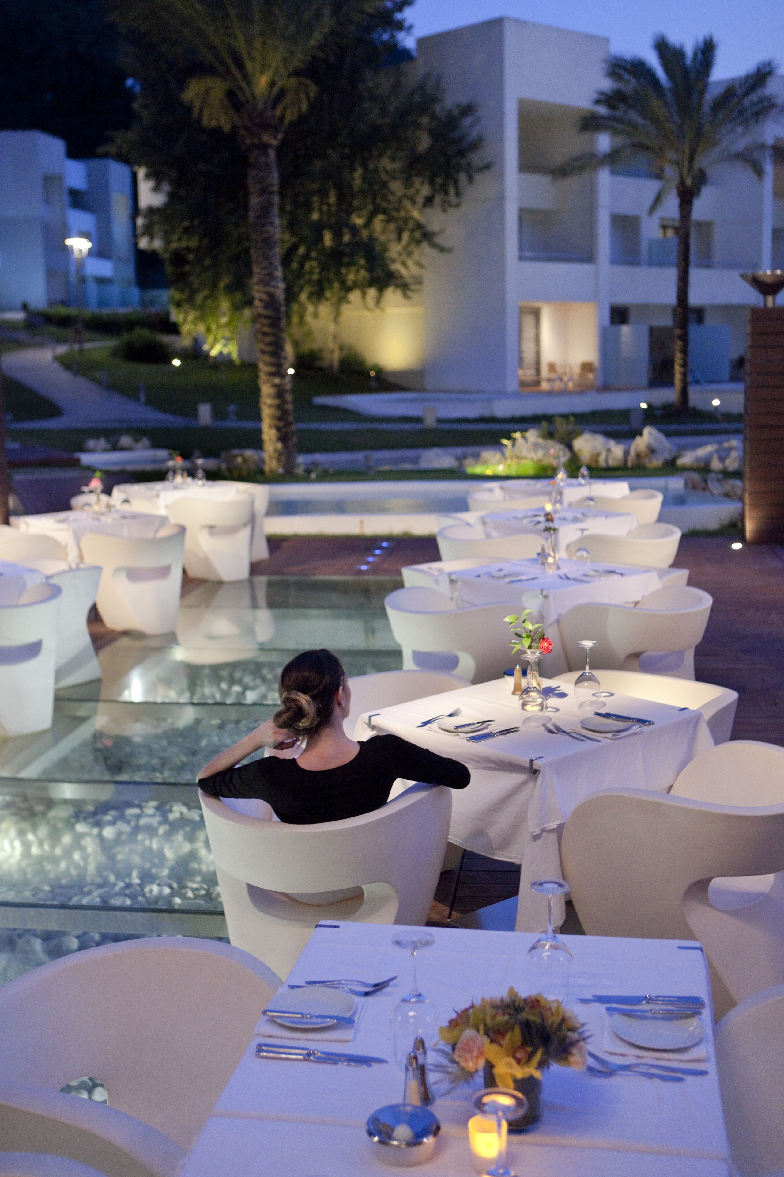 A dining area with round tables, white chairs, and a glass barrier. Tables are set with plates, cutlery, and candles.