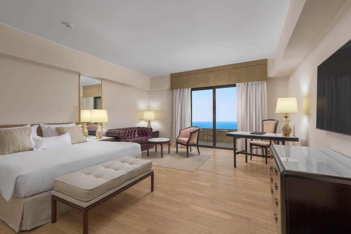 A hotel room with a large bed, a sitting area with a sofa and chairs, a small table, and a view of the ocean through a large window.