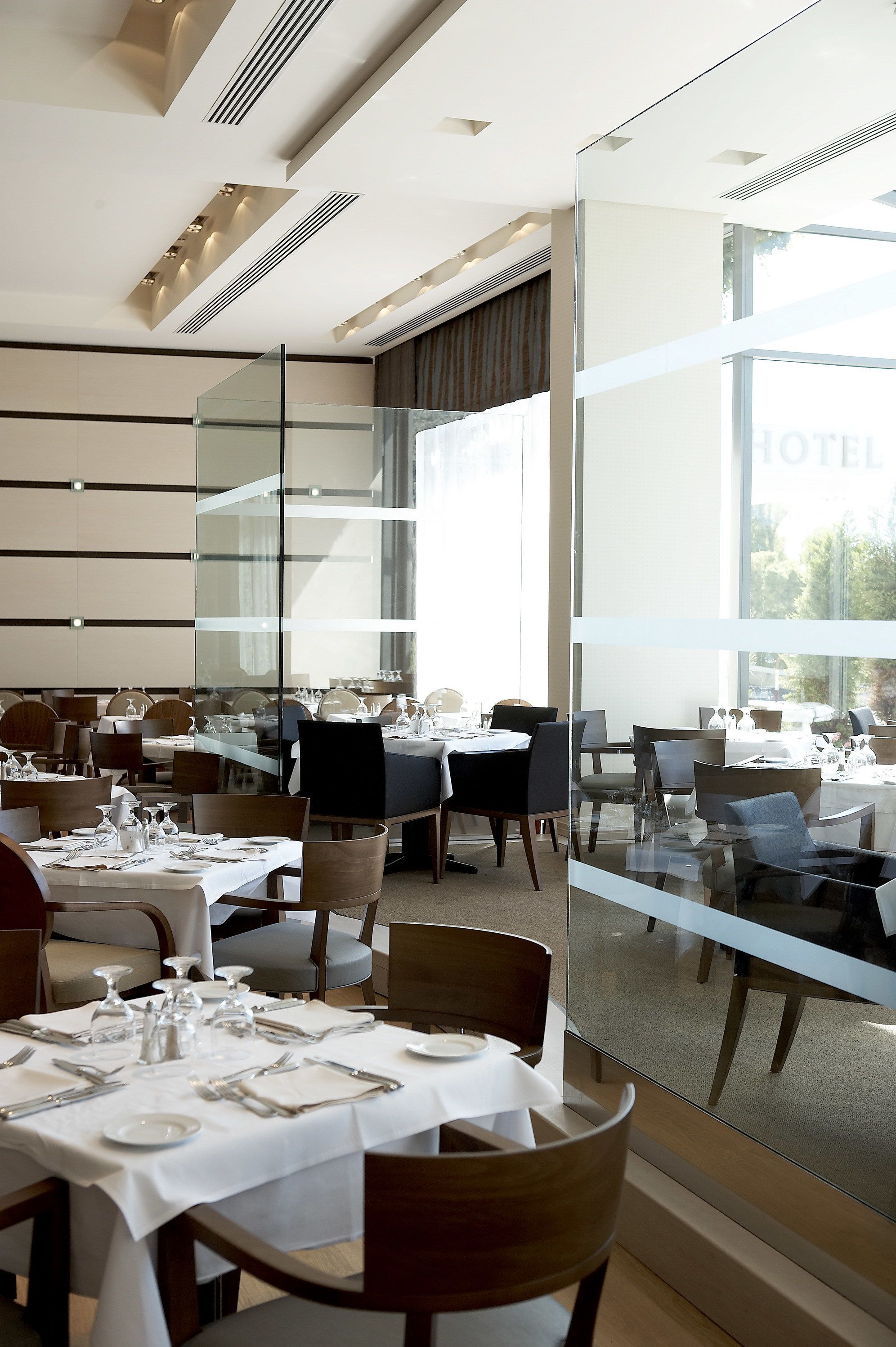 Un comedor de hotel moderno con varias mesas dispuestas para los huéspedes, amplias ventanas que ofrecen vistas y un panel de vidrio que separa el comedor del resto del hotel.