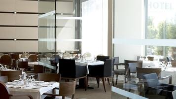 Un comedor de hotel moderno con varias mesas dispuestas para los huéspedes, amplias ventanas que ofrecen vistas y un panel de vidrio que separa el comedor del resto del hotel.