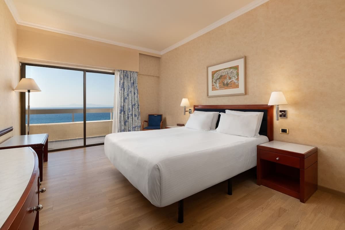 A hotel room with a large bed, a desk, a chair, a nightstand, and a view of the sea.