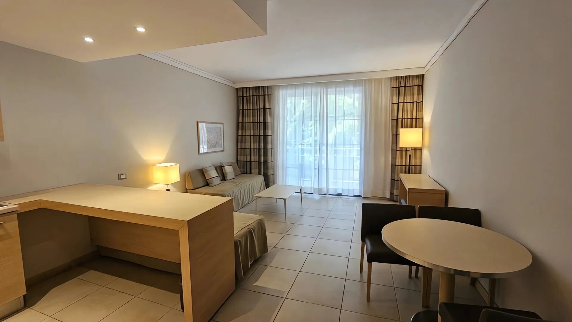 Una habitación moderna y luminosa con escritorio, sofá, mesa de comedor y un amplio ventanal con cortinas.