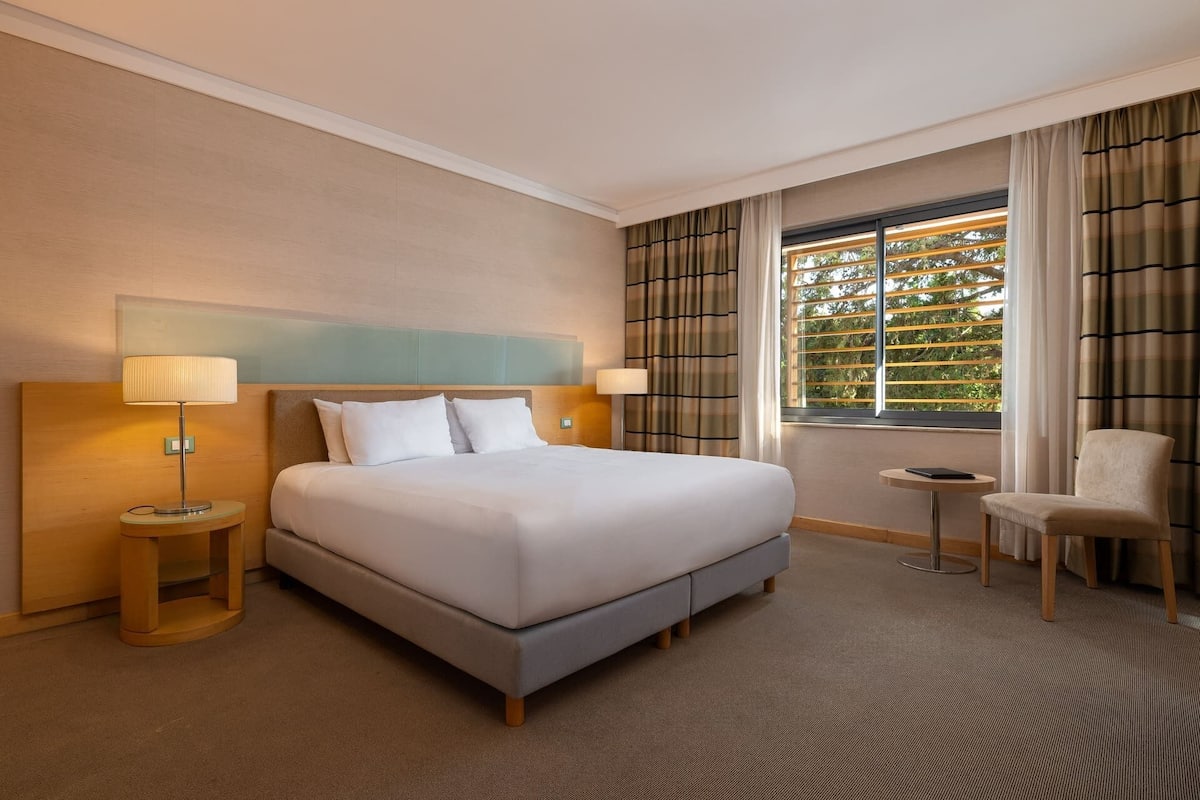 A hotel room with a large bed, two bedside lamps, a small table, a chair, and a window with curtains.