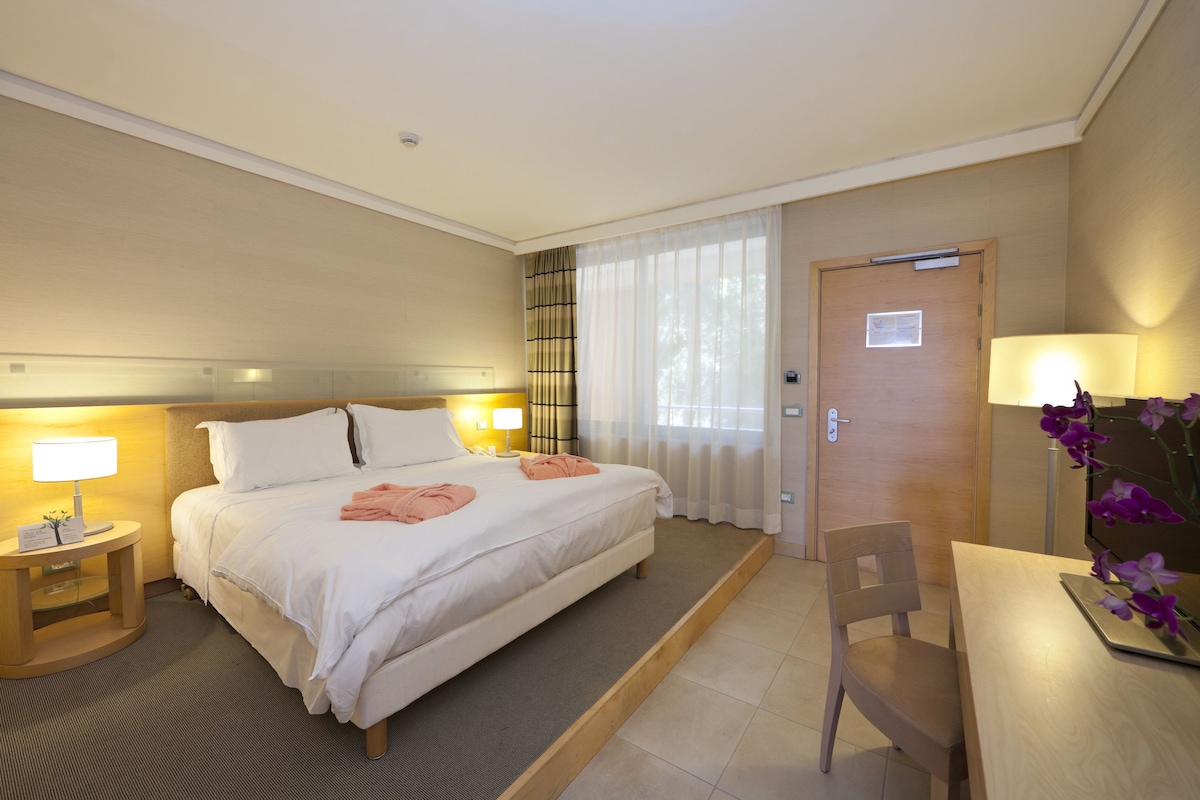 Habitación de hotel con cama, lámparas de noche, una silla de madera y un escritorio con lámpara.