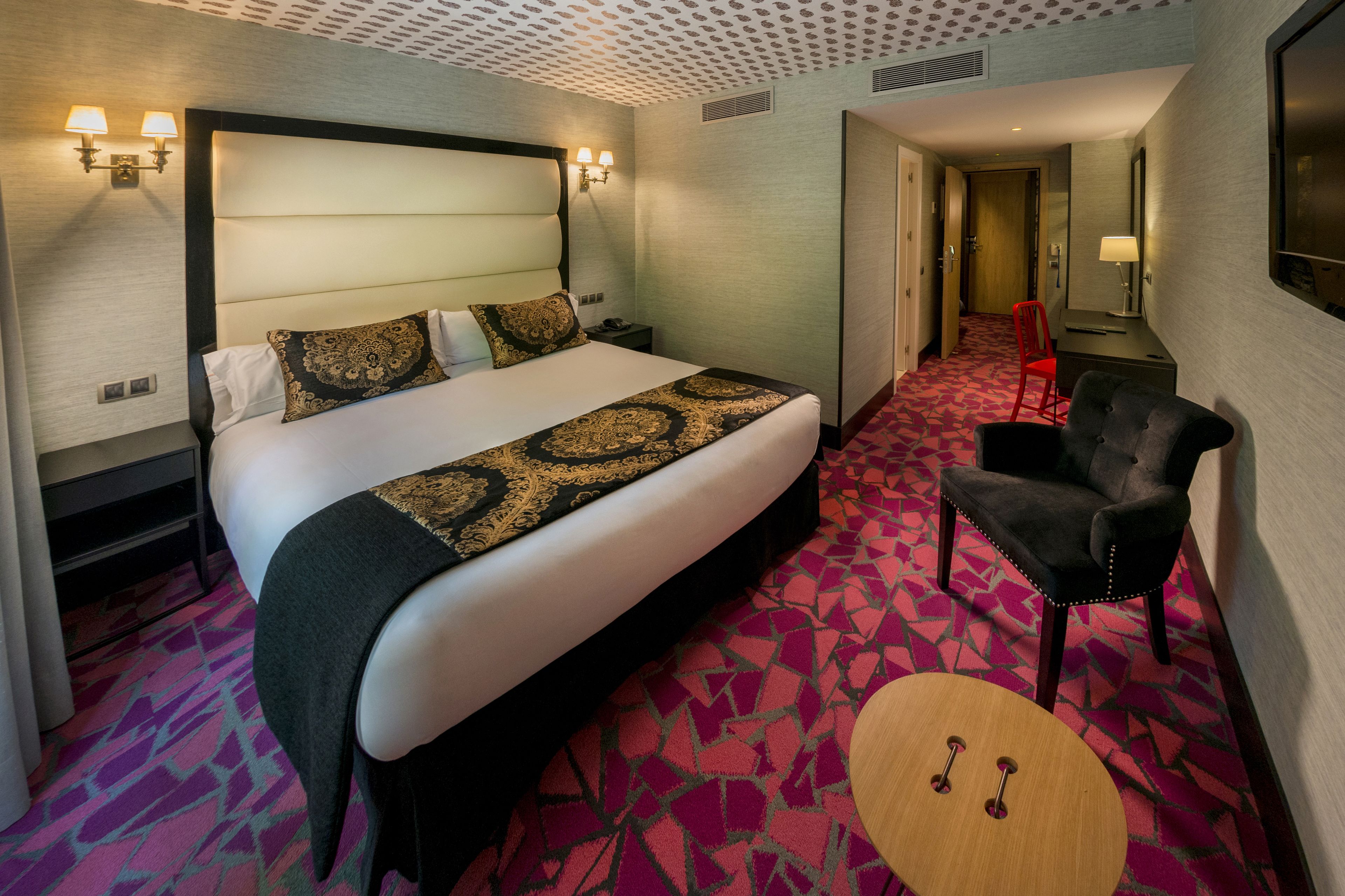 A hotel room with a large bed, a small round table, a red chair, and a nightstand.