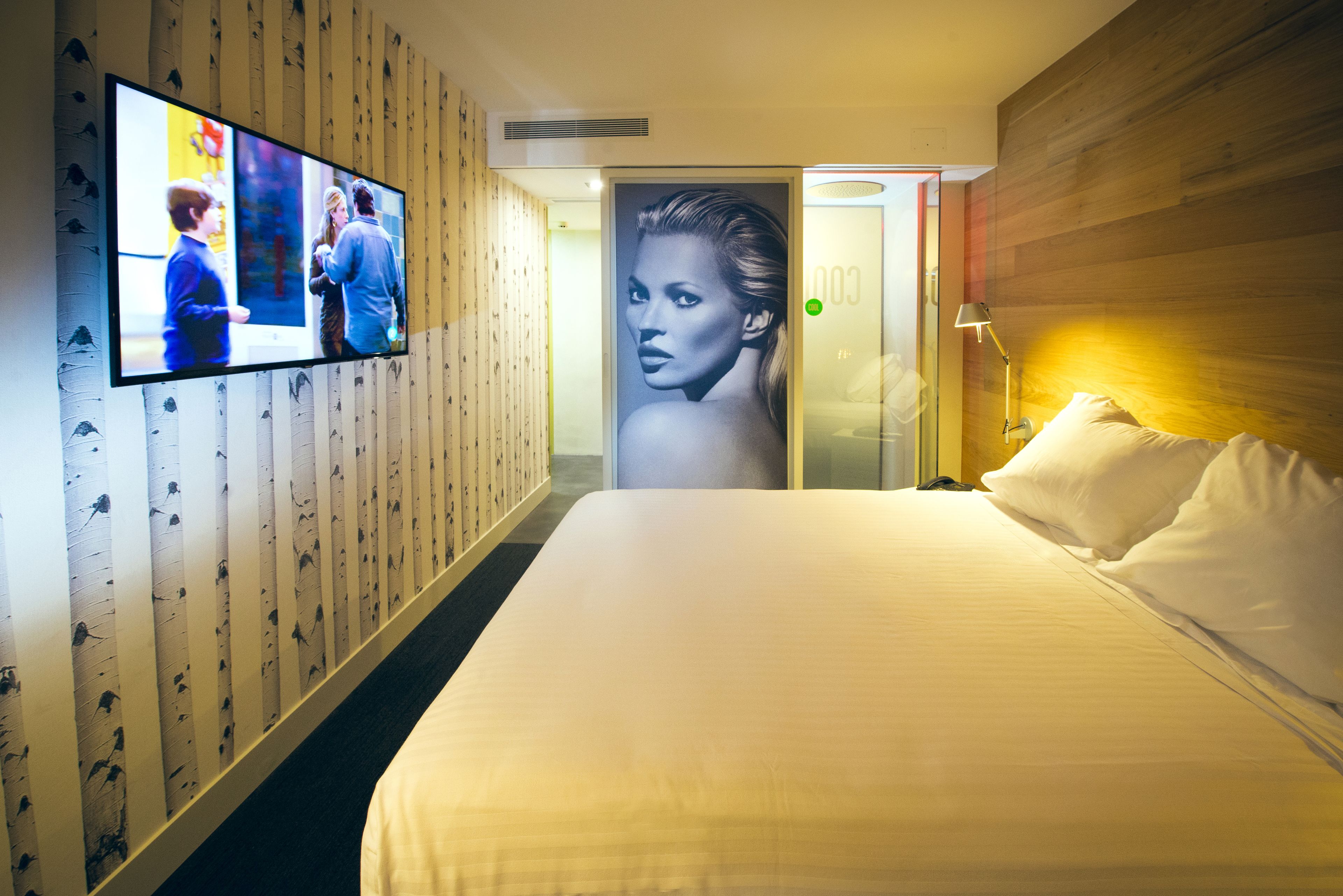 Habitación de hotel con una cama grande, un televisor montado en la pared y un cartel con la imagen de una mujer en la puerta.