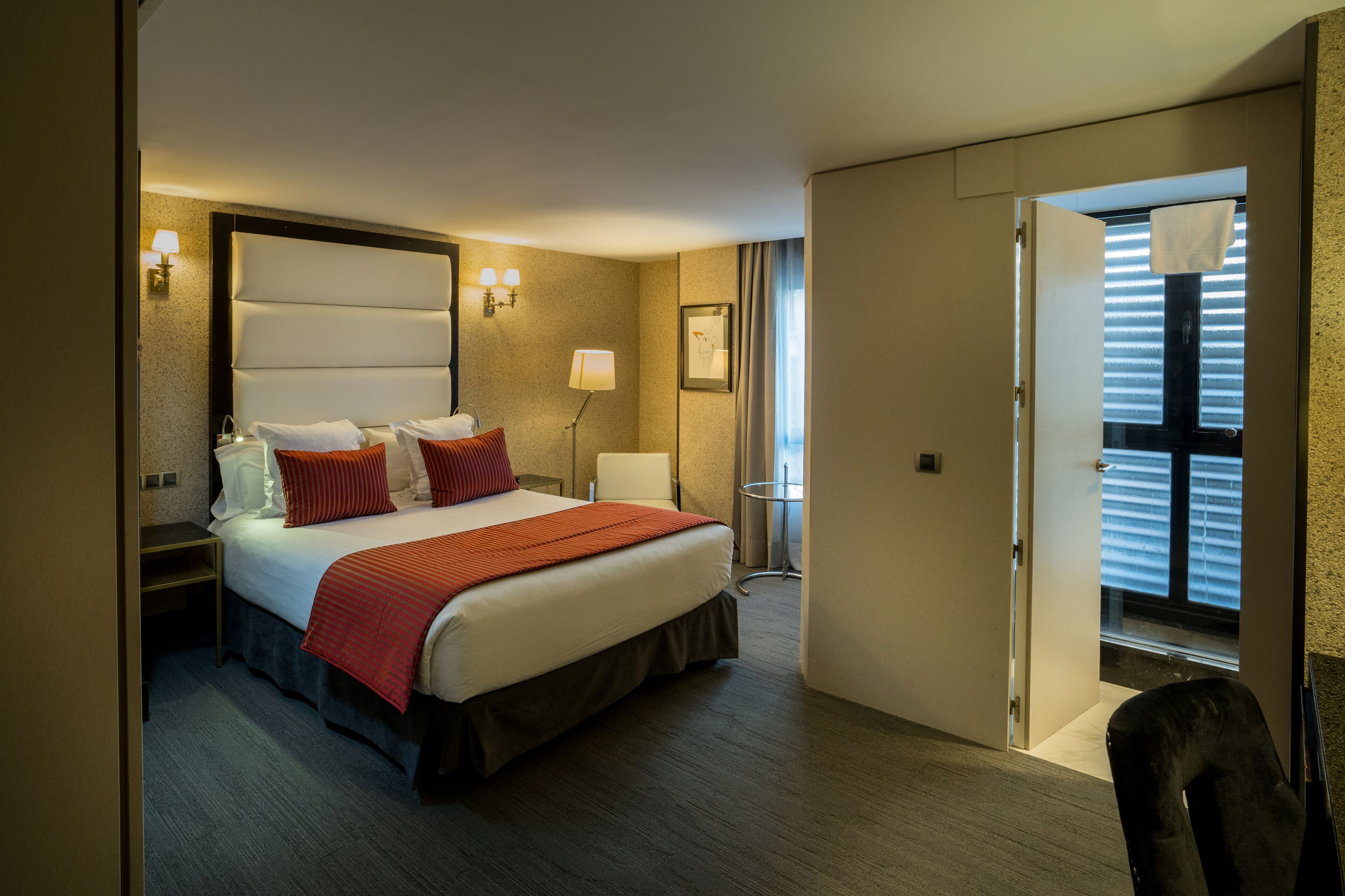 A hotel room with a bed, bedside tables, a lamp, a chair, and a window with blinds.