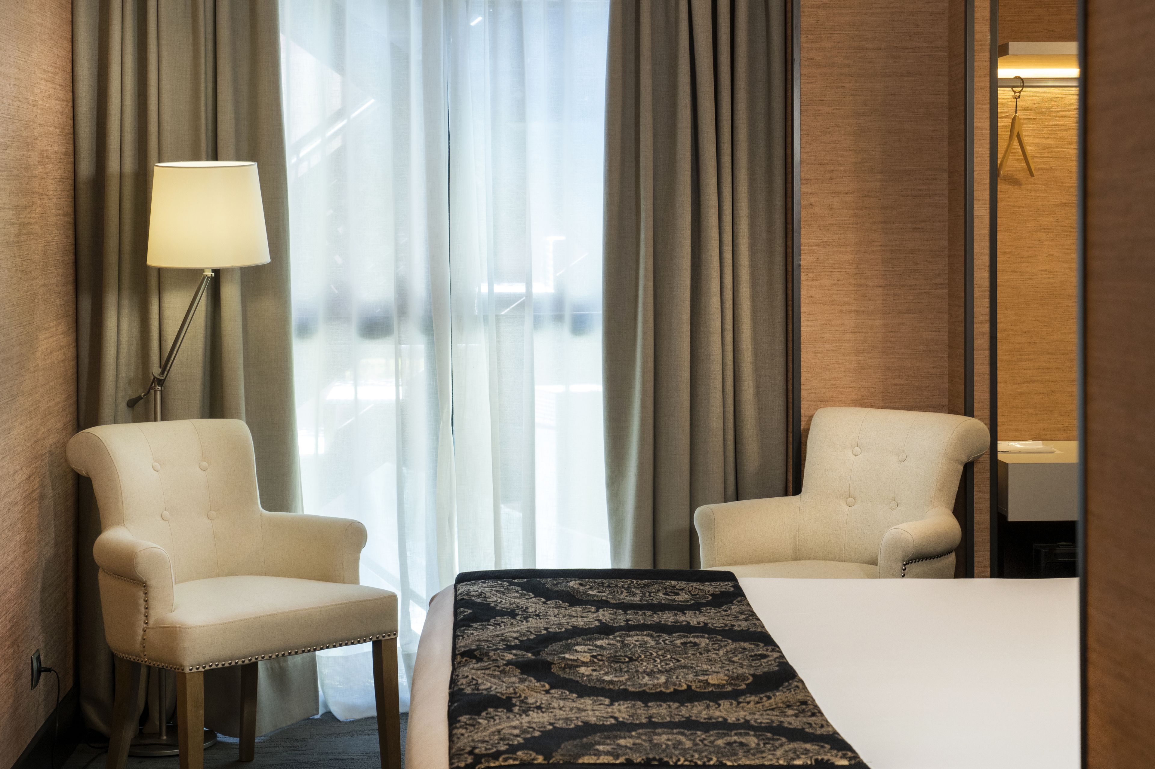 A hotel room with a bed, two armchairs, a lamp, and a view of the outside through large windows.