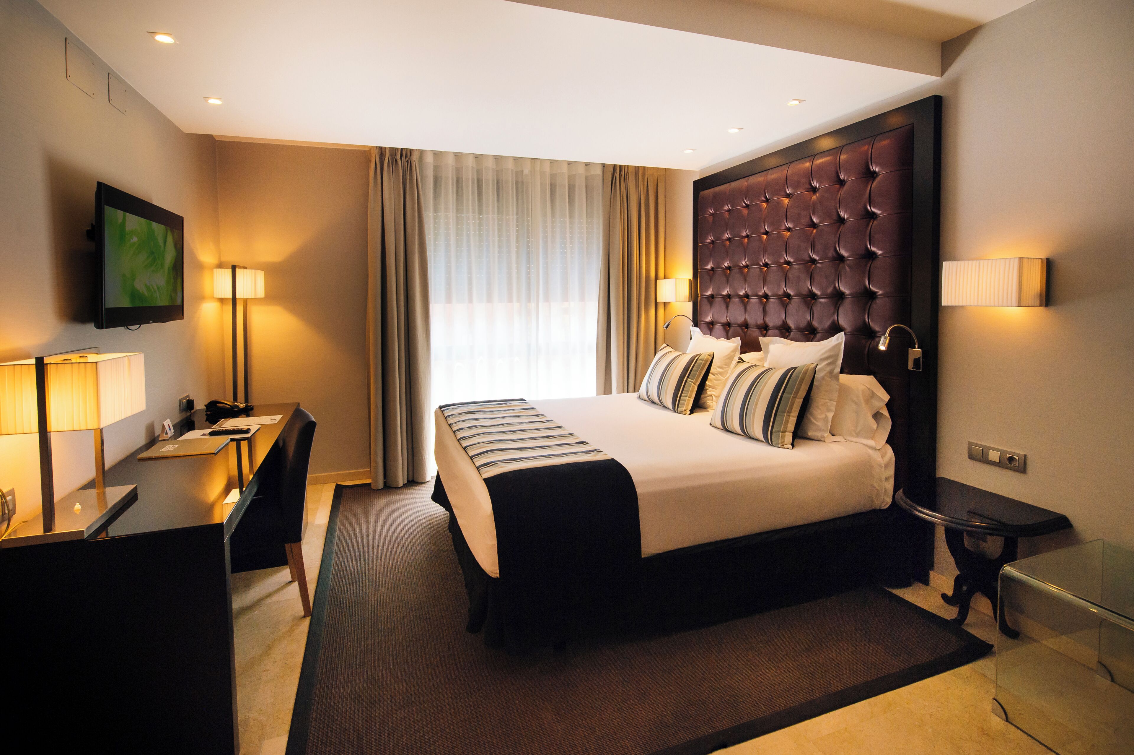 A hotel room with a bed, desk, chair, TV, and lamps.