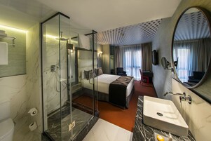 Ein Hotelzimmer mit einer gläsernen Dusche, einem Bett mit Kopfteil, einer Sitzecke mit Sessel und Tisch sowie einem großen Spiegel.