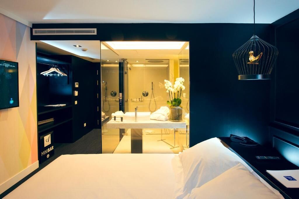 A hotel room with a bed, a bedside table with a lamp, a mirror, a minibar, and a hanging light fixture.