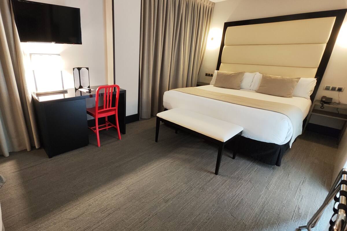 A hotel room with a bed, a desk, a chair, a bench, a TV mounted on the wall, and curtains.