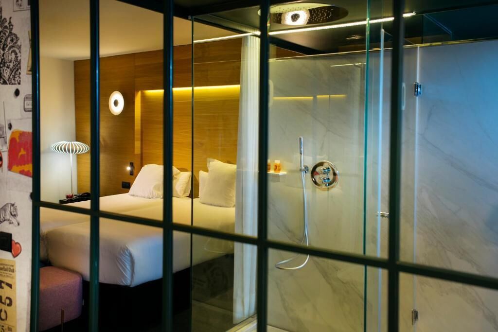 A hotel room with a glass partition, a bed, a shower, and a wall-mounted lamp.