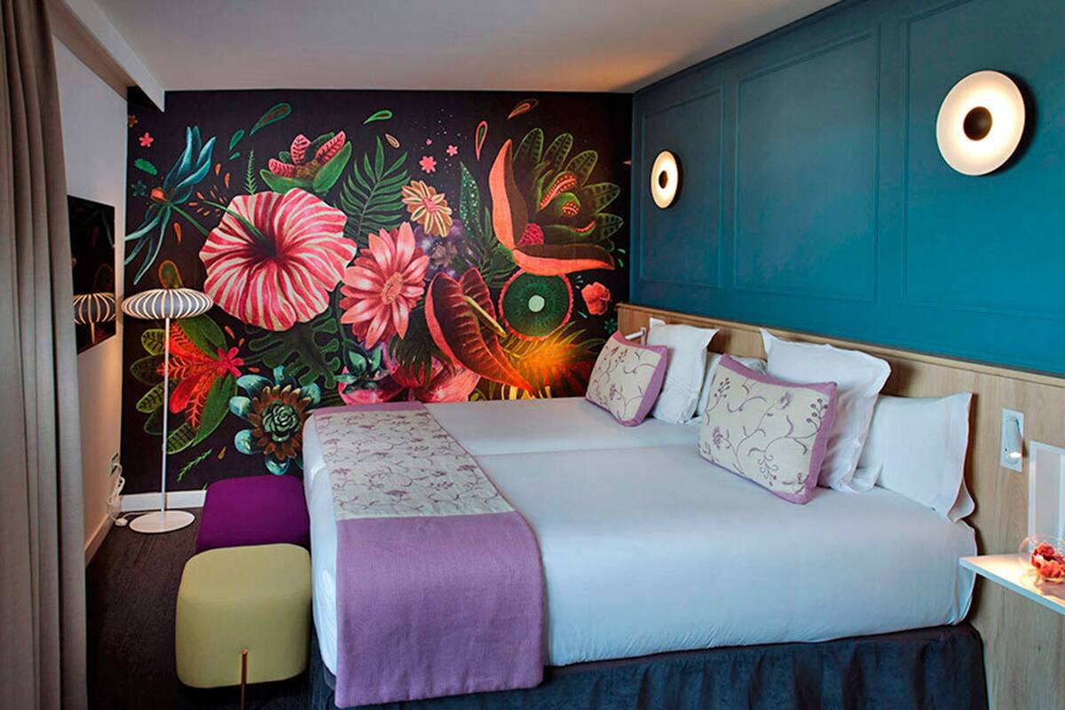A hotel room with a large bed, a colorful floral mural, a purple ottoman, and a small table with a lamp.