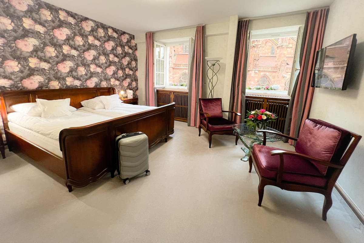 A hotel room with a large bed, a desk with a chair, a suitcase, and a view of a building through the window.