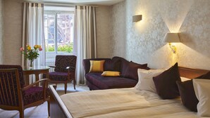Una habitación de hotel con cama, sofá, sillón, una mesa con flores y una ventana con cortinas.