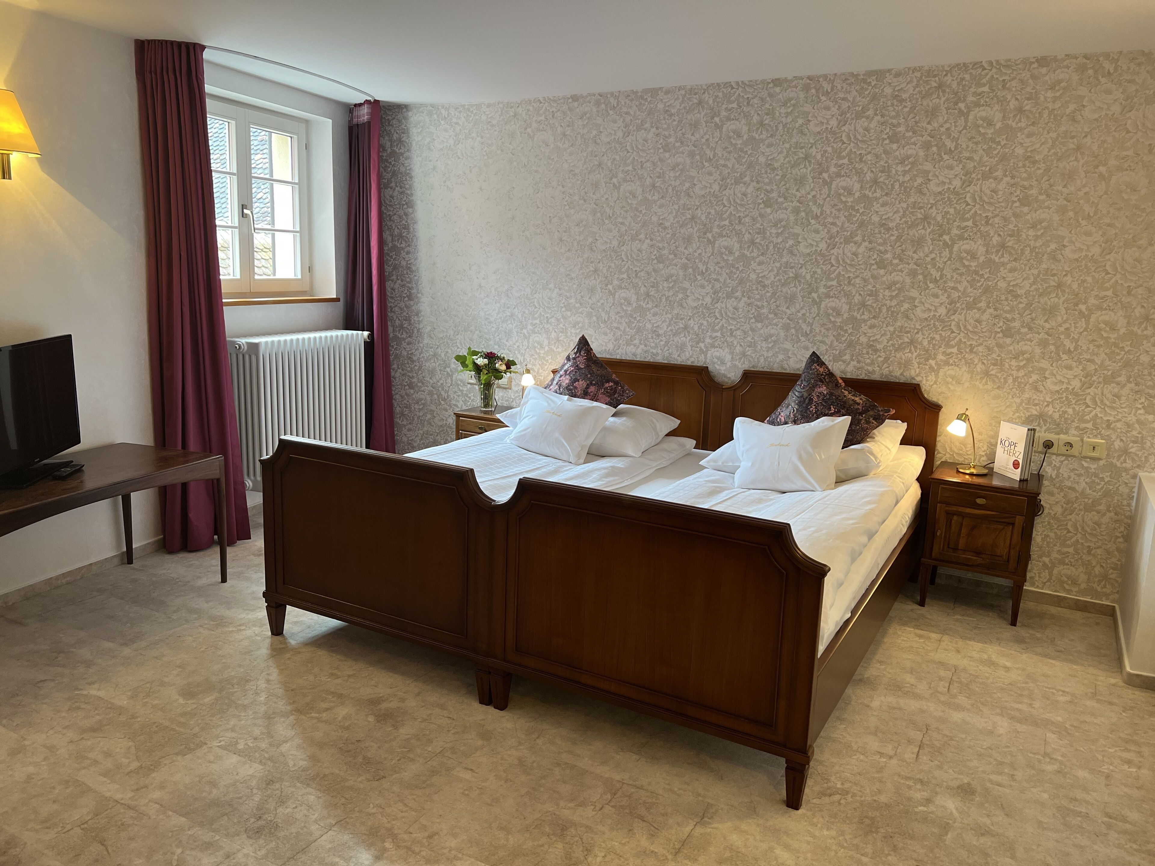 A neatly arranged bedroom with a wooden bed, a bedside table with a lamp, a nightstand, and a television on a desk.