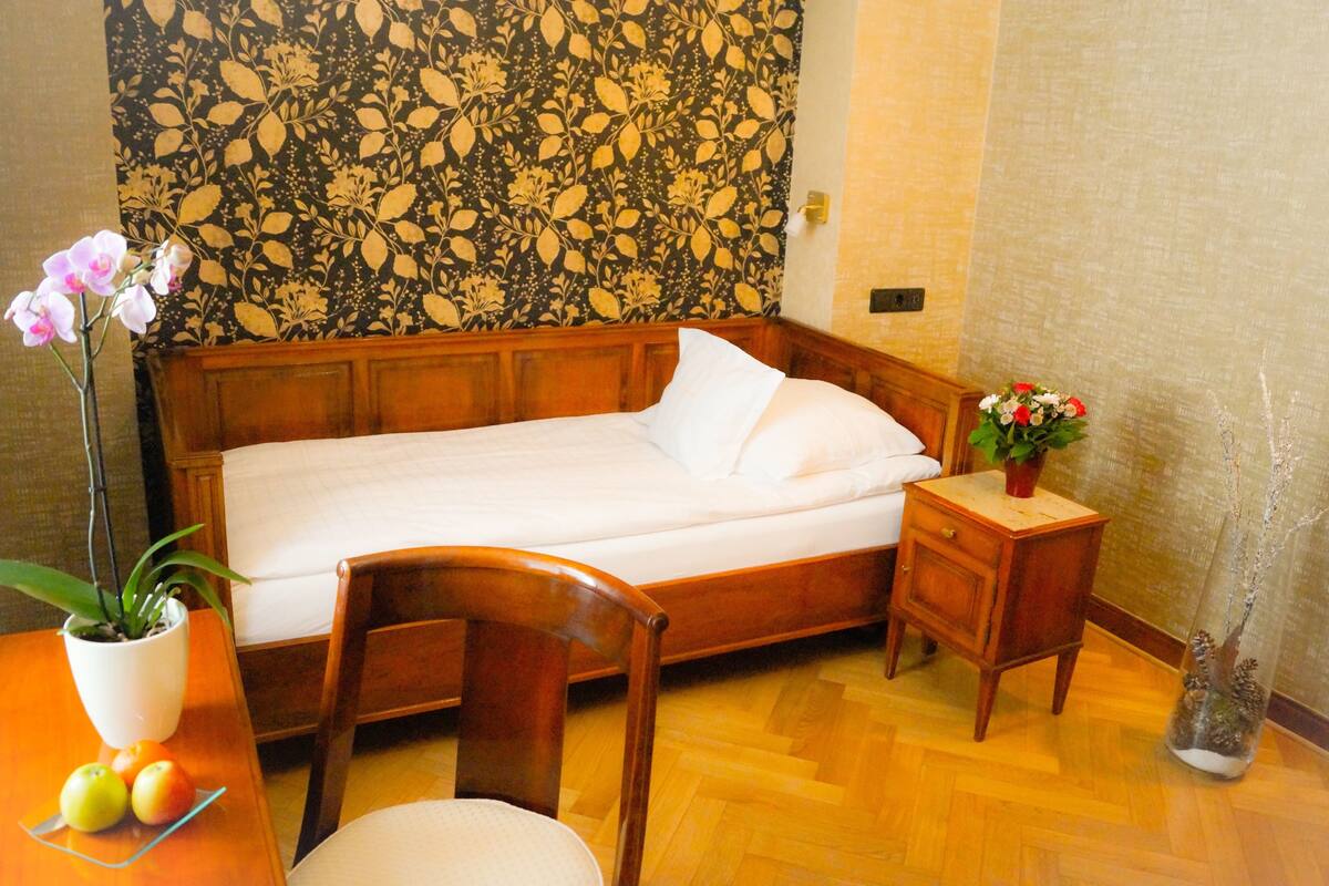 A neatly made bed with a wooden headboard, a small bedside table with a vase of flowers, and a chair. There is a potted plant on a table next to the bed.