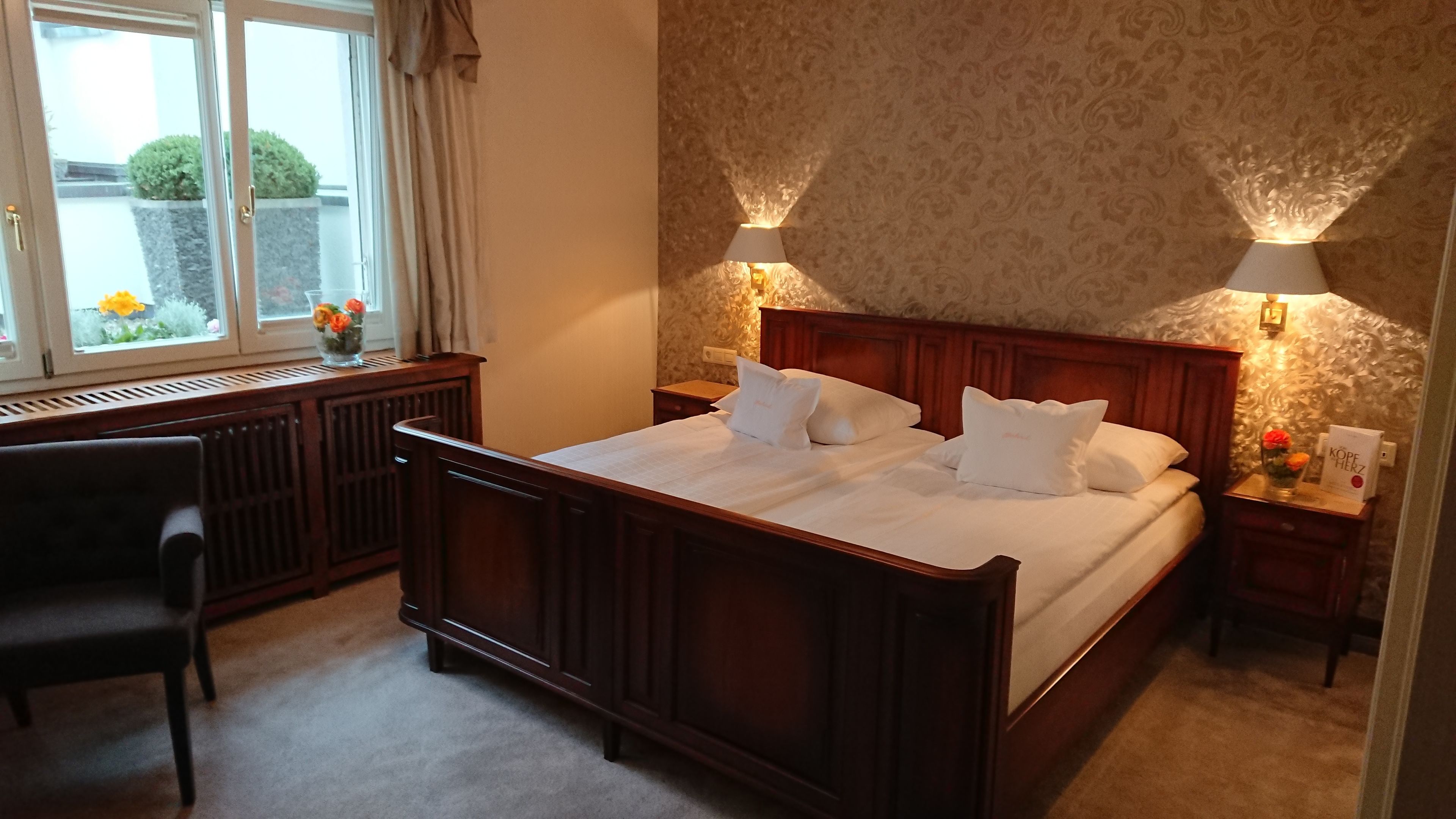 A bedroom with a large bed, a desk, and a chair. There is a window with a view of a garden.