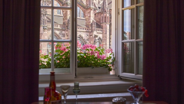 Una mesa con una botella, copas de vino y un plato de fruta junto a una ventana que ofrece vistas a un edificio histórico y flores.