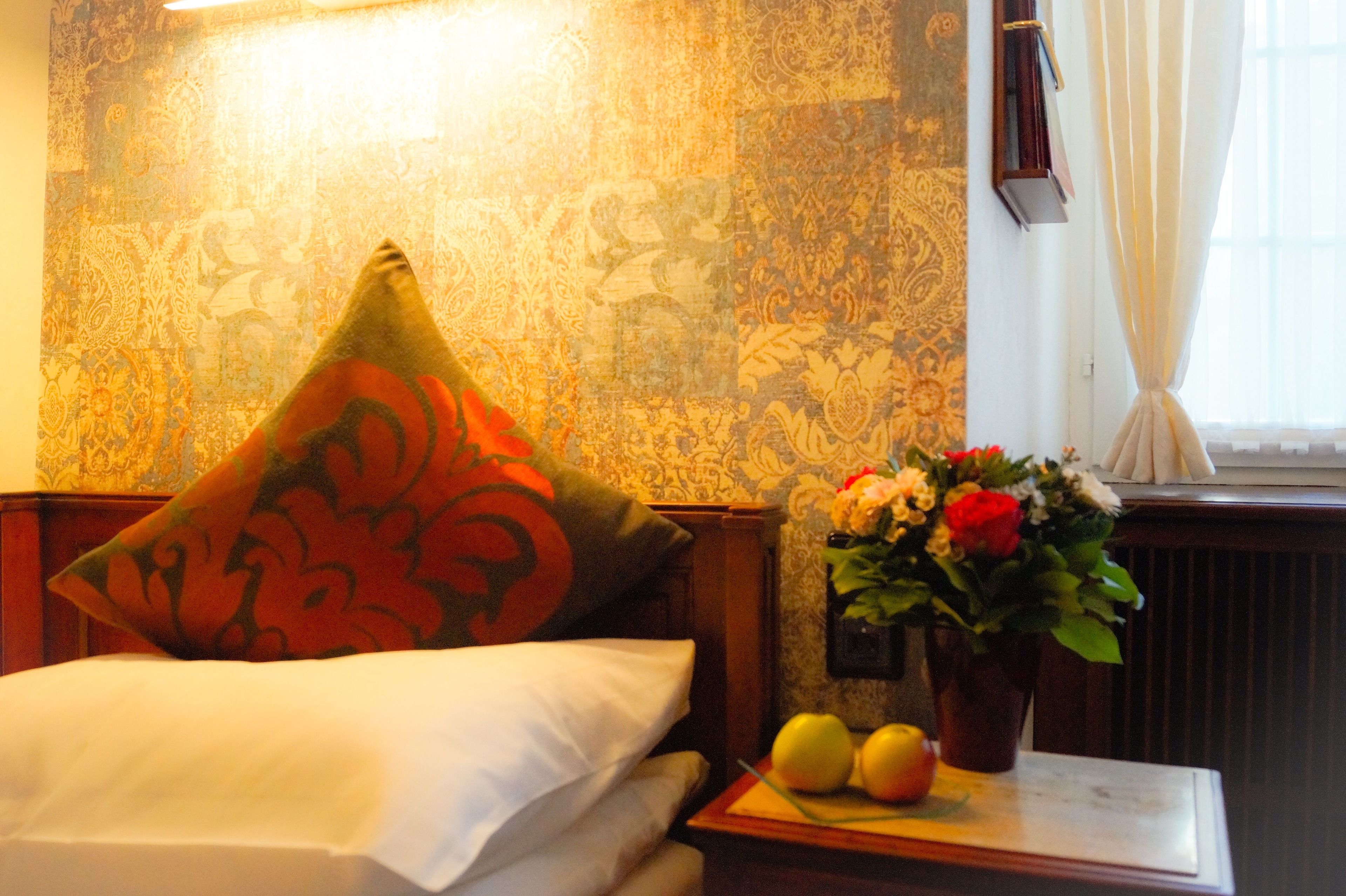 Una camera d'albergo con carta da parati a motivi, un letto con cuscini bianchi e arancioni, un comodino con un vaso di fiori e delle mele, e una finestra con tende.