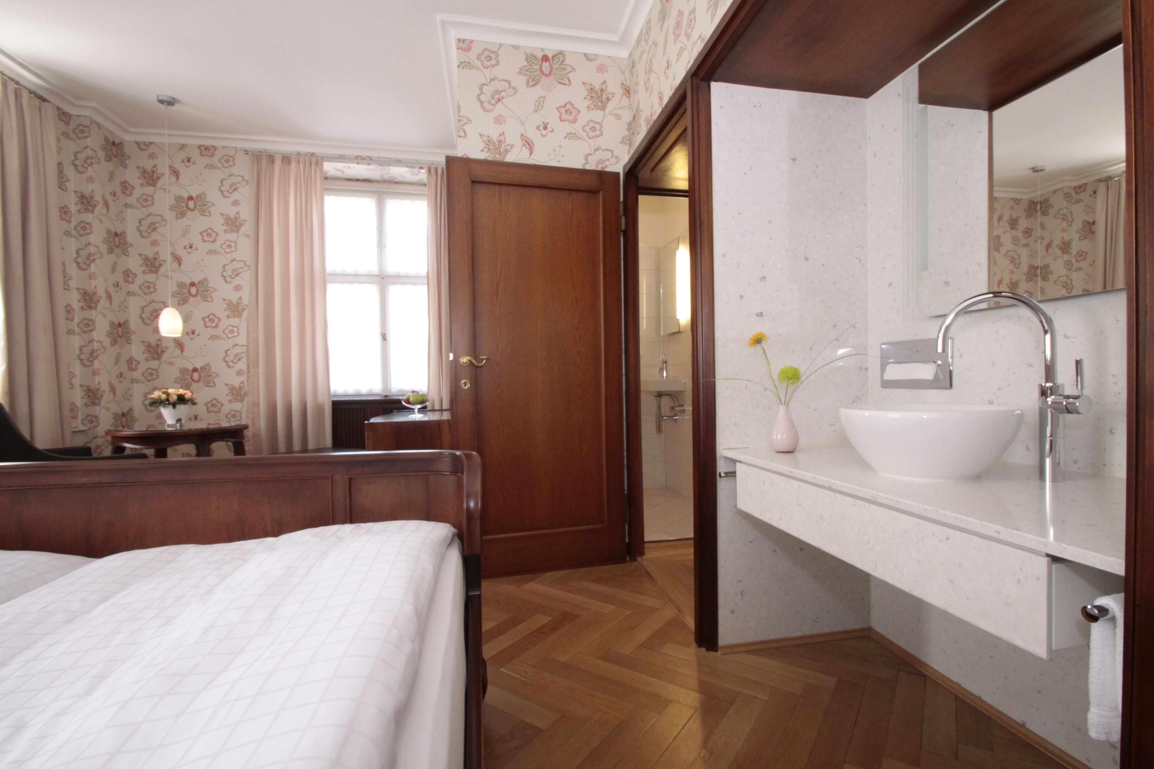 A hotel room with a wooden bed, a bathroom with a white sink and mirror, and a door leading to another room.