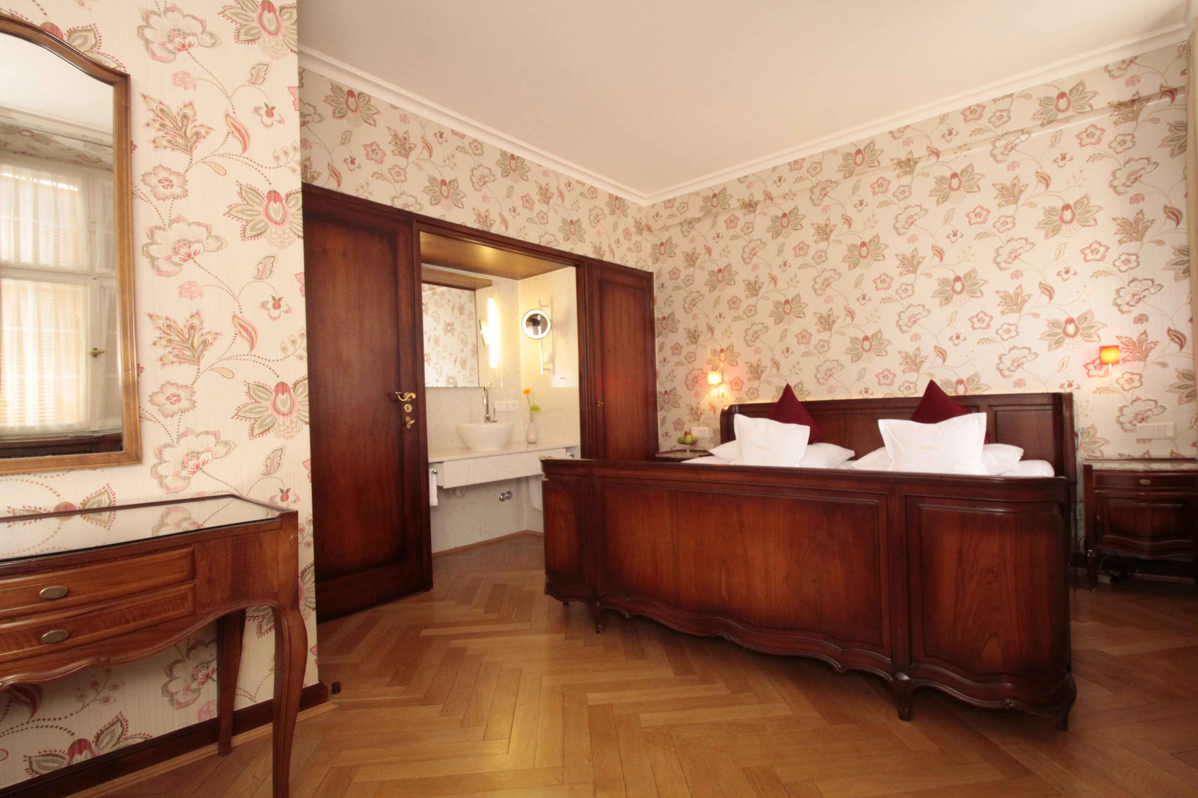 Ein Schlafzimmer mit einem Holzbett, einem Schminktisch, einem Spiegel und einem durch eine geöffnete Tür einsehbaren Badezimmer.