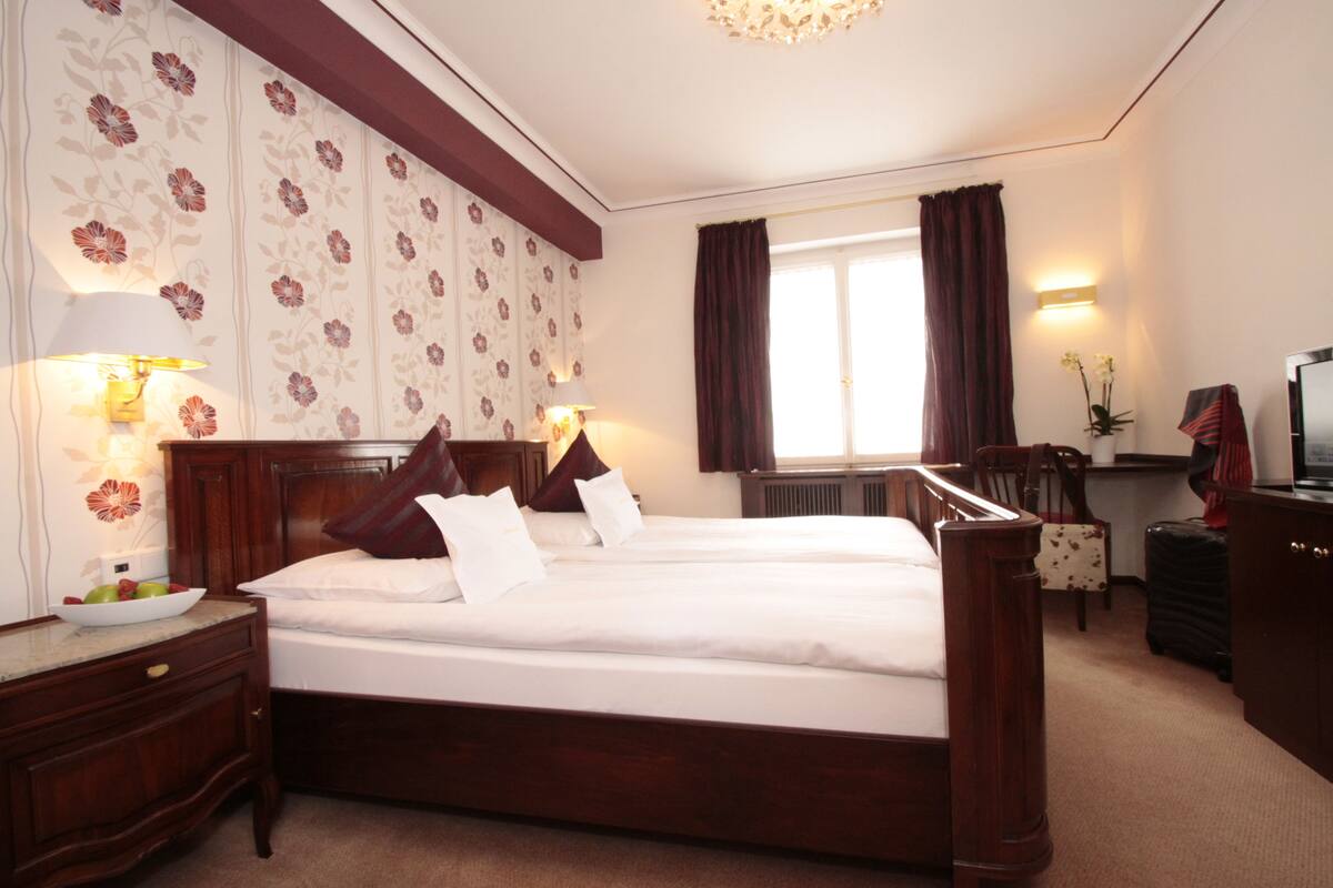 A hotel room with a large bed, wooden headboard, two bedside lamps, a dresser, a desk, and a window with curtains.