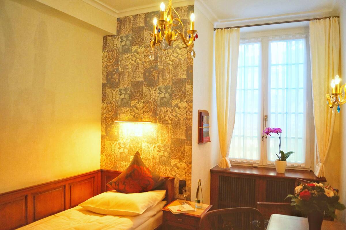 A hotel room with a bed, a nightstand, a lamp, a window with curtains, and a potted plant.