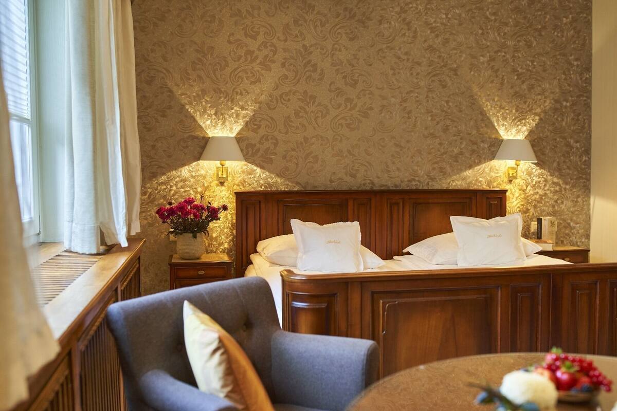 A hotel room with a wooden bed, two bedside lamps, a nightstand with a vase of flowers, a chair, and a round table with fruit.
