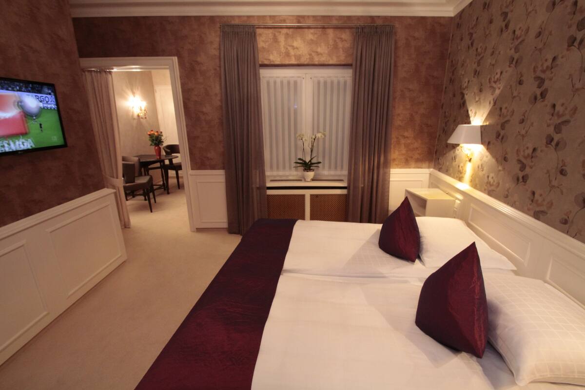 A hotel room with a large bed, two red pillows, a TV on the wall, a desk with a chair, and a window with curtains.