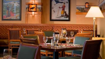 Un ristorante con arredi in legno, un tavolo apparecchiato con bicchieri e posate, e manifesti alle pareti raffiguranti scene di sci.