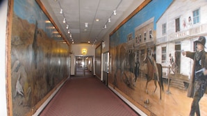 Un couloir avec une moquette rouge à motifs, de grandes fresques représentant des scènes historiques et une enseigne « Dodge House » sur le mur de droite.