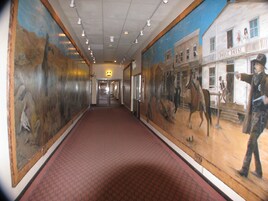 Un pasillo con alfombra roja de estampado, grandes murales que representan escenas históricas y un letrero de 'Dodge House' en la pared derecha.