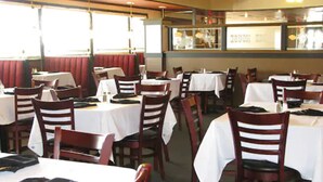 L’intérieur d’un restaurant, avec des banquettes et des tables, dont certaines sont recouvertes de nappes blanches, et des chaises en bois.