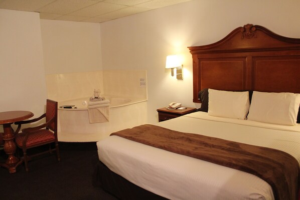 A hotel room with a large bed, a wooden headboard, a bedside table with a lamp, a chair, and a small table with a telephone.