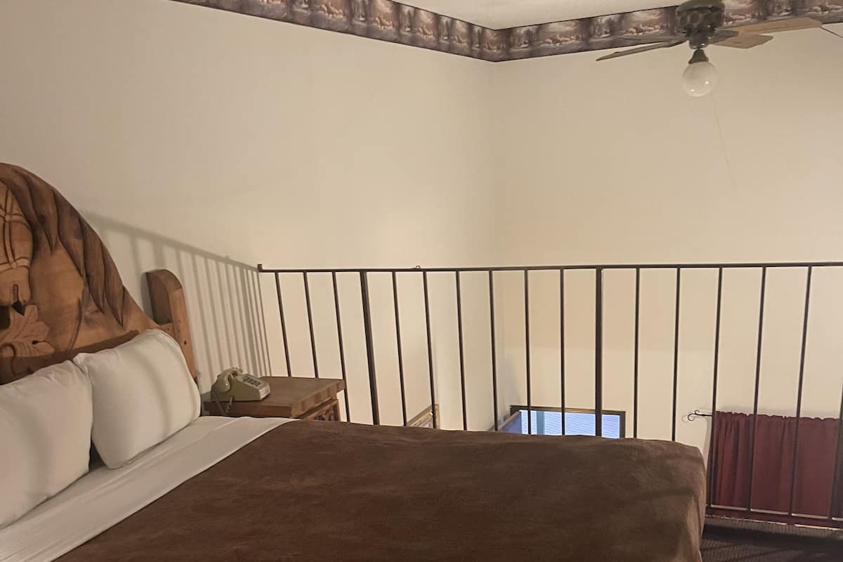 Une chambre à coucher avec un grand lit, une tête de lit en bois, une table de chevet, un téléphone, un ventilateur de plafond et une fenêtre avec des rideaux.