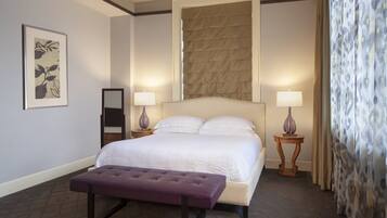 Una camera d'albergo con un letto grande, due lampade sui comodini, una testiera, un tavolino, una panca, un quadro appeso alla parete e una finestra con persiane.