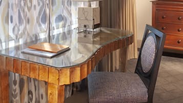 A modern hotel room with a glass desk, a chair, a lamp, and a wooden dresser.