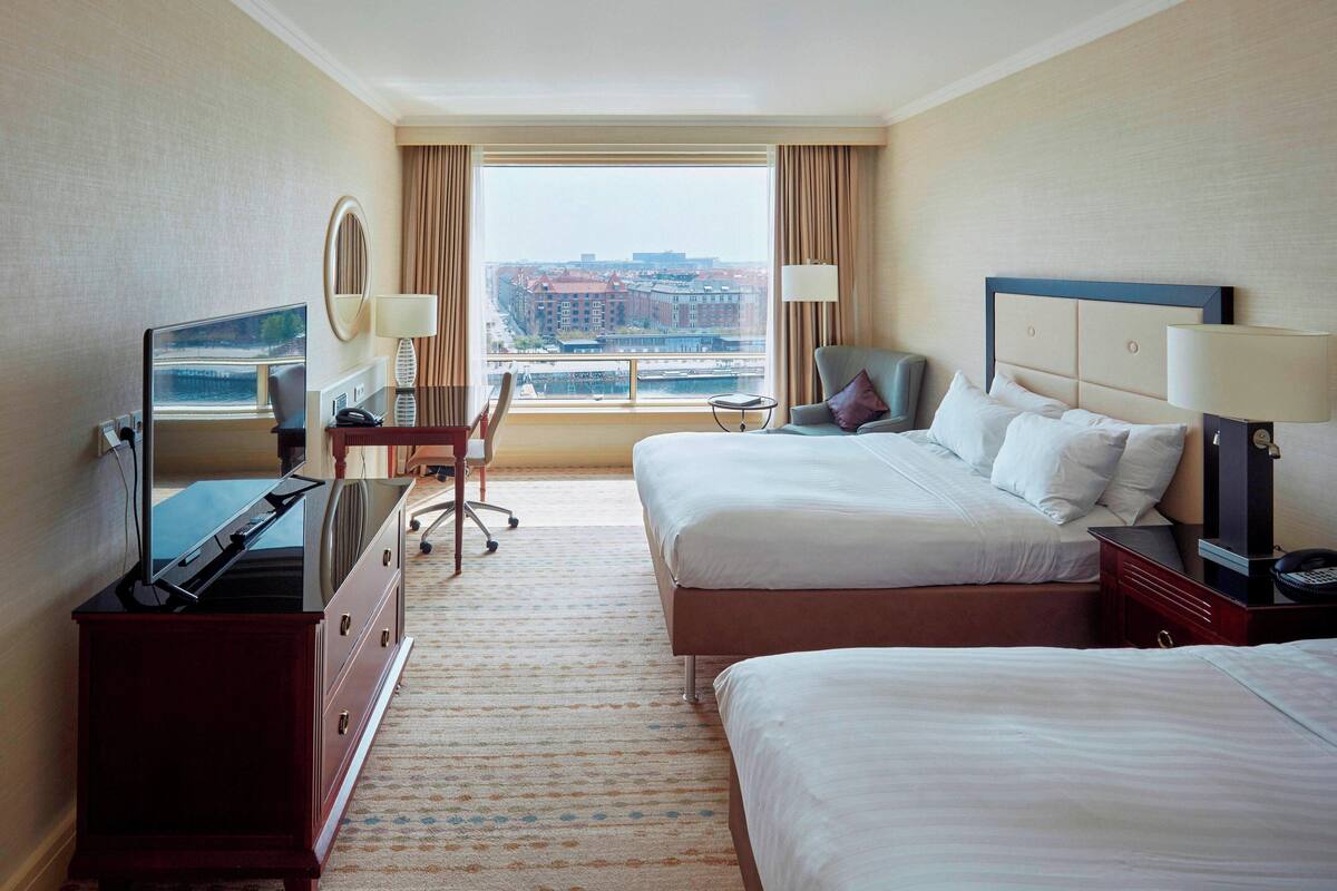 Habitación de hotel con una cama grande, una cama más pequeña, un escritorio con silla, televisor y vista a la ciudad a través de un ventanal amplio.
