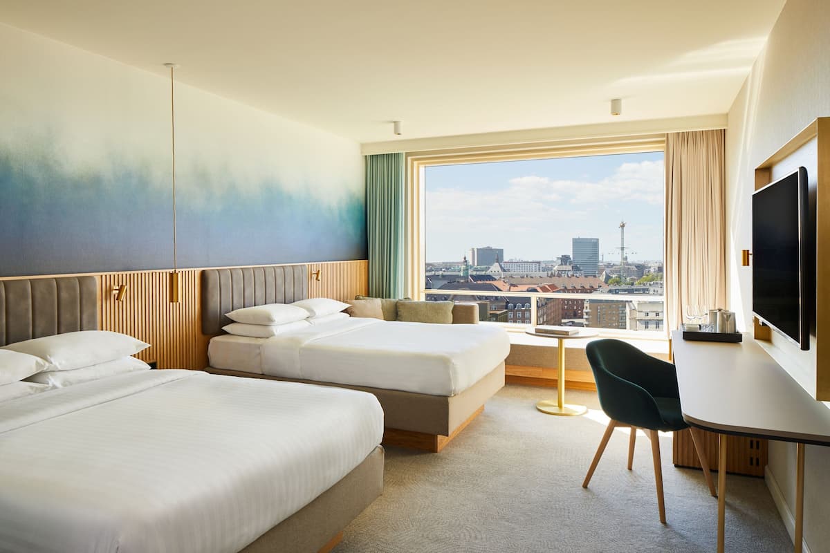 Habitación de hotel con dos camas, un escritorio, una silla y un ventanal con vistas a la ciudad.