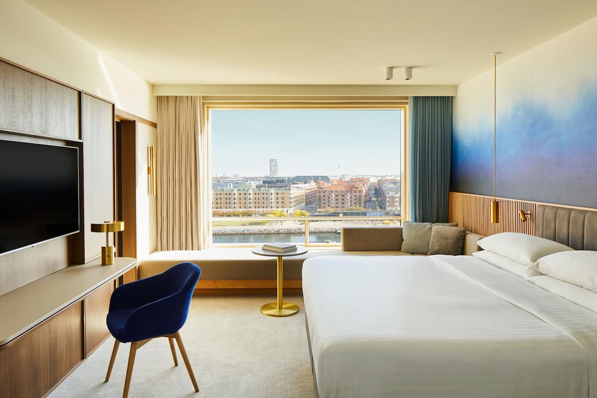 Una habitación de hotel con una cama grande, televisión, escritorio y vistas a un paisaje urbano a través de un ventanal.