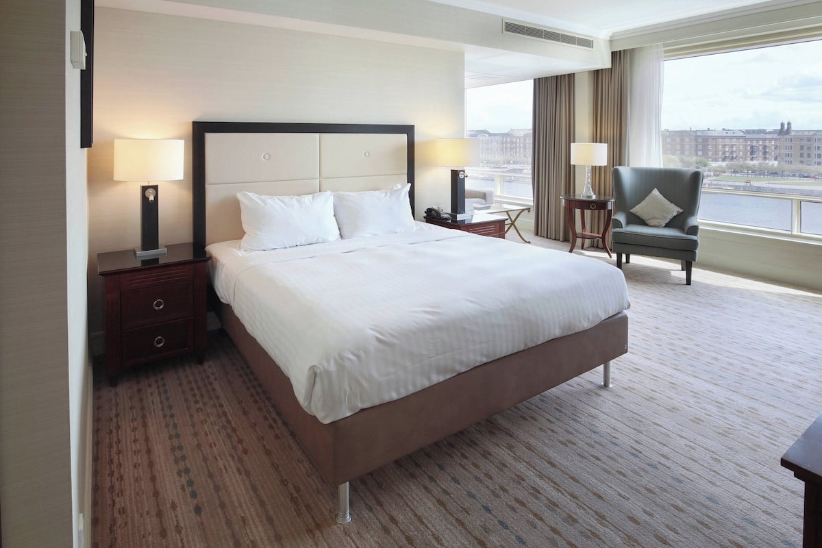 Habitación de hotel con una cama grande, dos mesitas de noche con lámparas, una silla y vistas a un paisaje urbano a través de un gran ventanal.